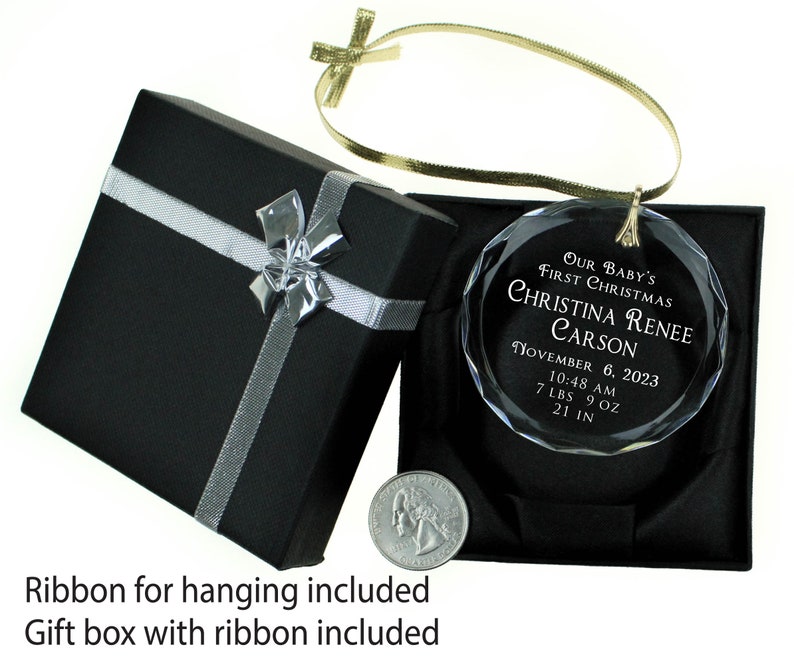 May include: A clear glass Christmas ornament with a gold ribbon. The ornament is engraved with "Our Baby's First Christmas Christina Renee Carson November 6, 2023 10:48 AM 7 LBS 9 OZ 21 IN". The ornament is in a black gift box with a silver ribbon.