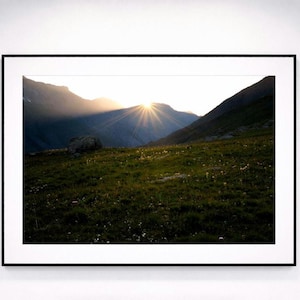 Peut inclure: Photographie encadrée d'un paysage de montagne au coucher du soleil. Les rayons du soleil percent les sommets, illuminant le premier plan herbeux. L'image est encadrée dans un cadre noir, créant un contraste saisissant.