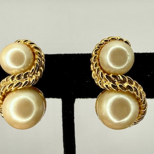 May include: A pair of gold-toned earrings featuring two large, cream-colored, round faux pearls. The pearls are connected by a twisted, rope-like gold metal design. The earrings are displayed against a black background.