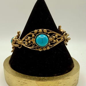 May include: A gold-toned bracelet with a filigree design, featuring three large, round turquoise stones. The bracelet is displayed on a black velvet cone, set on a gold-painted wooden base. The intricate metalwork is accented with small, decorative details.