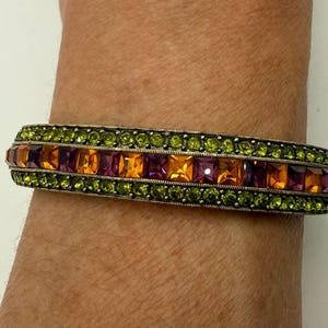 May include: A hinged bangle bracelet featuring a central row of square-cut orange and purple gemstones flanked by two rows of small, round, lime green crystals set in a metallic frame with a textured, darkened finish.