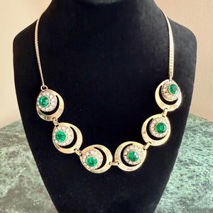May include: A gold-coloured necklace with emerald green and clear crystal accents. The necklace features circular pendants with a central green stone surrounded by clear crystals. It is displayed on a black mannequin.