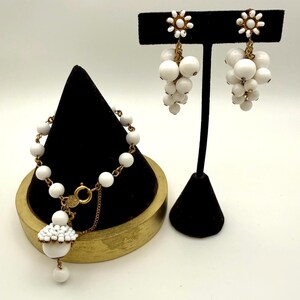 May include: A jewelry set consisting of a bracelet and a pair of drop earrings. The bracelet features white beads on a gold-tone chain with a central cluster charm. The earrings have a white flower-shaped top with a cluster of white beads.