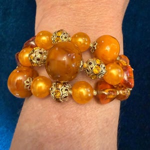 May include: A multi-strand bracelet featuring large, smooth, amber-colored resin beads interspersed with smaller, intricate gold-tone metal filigree beads. The design includes a central, oversized round bead, creating a layered, textured appearance.