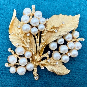 May include: A gold-toned brooch in the shape of a leaf and pearl cluster. The brooch features a textured leaf design with a cluster of white pearls and small, sparkling crystals. The brooch is set against a teal background.