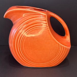 May include: A vibrant orange ceramic pitcher with a curved spout and a large handle. The pitcher features concentric, ridged detailing around the body. The design is a classic, retro style, perfect for serving beverages.