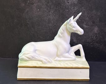 May include: A white ceramic unicorn figurine in a reclining position, featuring a single horn on its forehead and a flowing mane. The unicorn rests upon a rectangular base with a pale yellow top surface and a gold-painted trim along the edges.