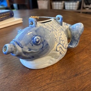 May include: A blue and white ceramic teapot shaped like a fish. The teapot has a spout, handle, and a metal wire handle. The fish design includes scales and decorative patterns. The teapot is sitting on a wooden table.
