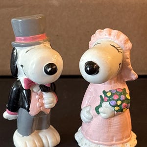 May include: Two ceramic figurines of Snoopy characters dressed for a wedding. The left figure wears a gray top hat with a pink band, a black jacket, and a pink vest. The right figure wears a pink dress and veil, holding a colorful floral bouquet.