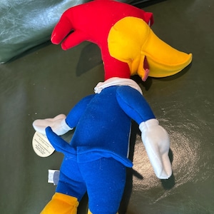 May include: A plush toy of Woody Woodpecker, featuring a red head, yellow beak, and blue outfit. The toy has white gloves and yellow feet. A tag is attached to the toy.
