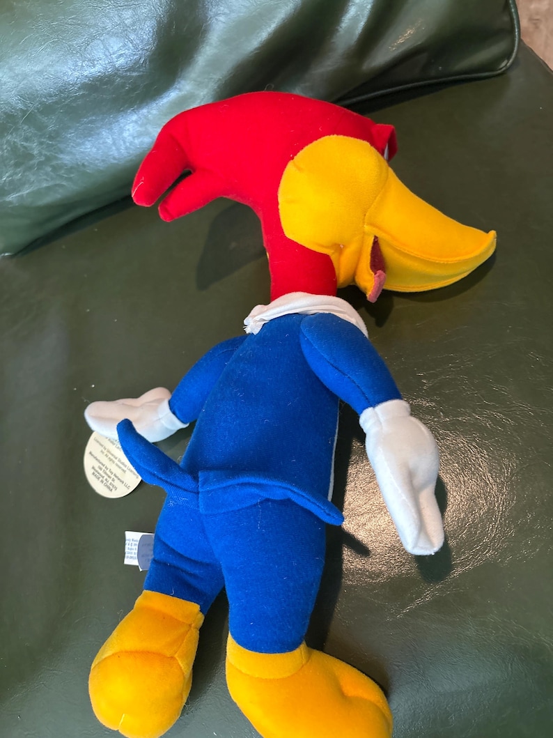 May include: A plush toy of Woody Woodpecker, featuring a red head, yellow beak, and blue outfit. The toy has white gloves and yellow feet. A tag is attached to the toy.