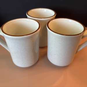 May include: Three ceramic mugs with a speckled off-white finish and a smooth brown rim. Each mug features a cylindrical body and a curved handle on the side. The interior of each mug is a solid off-white color, matching the exterior base tone.