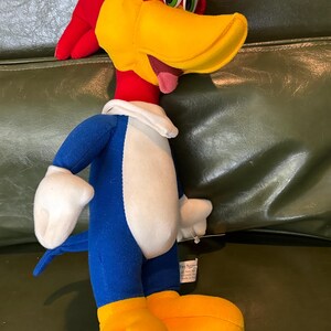 May include: A plush Woody Woodpecker doll. The cartoon character has a red head and beak, blue body, white chest and hands, and yellow feet. The doll has green eyes and a pink tongue. The doll is made of soft fabric.