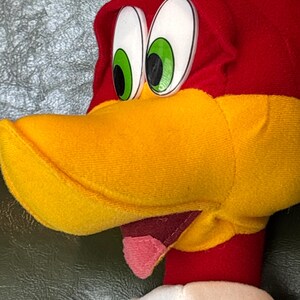 May include: Close-up of a plush Woody Woodpecker toy. The toy features a red head with a yellow beak, large white eyes with green irises, and a pink tongue. The toy has a white collar and a blue shirt.