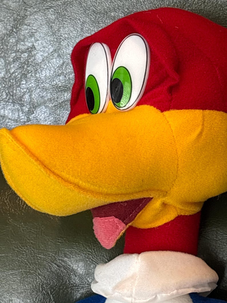 May include: Close-up of a plush Woody Woodpecker toy. The toy features a red head with a yellow beak, large white eyes with green irises, and a pink tongue. The toy has a white collar and a blue shirt.