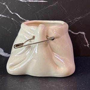May include: A cream-colored ceramic vessel shaped like a small, gathered fabric pouch. The top opening is cinched with a metallic safety pin that pierces through the ceramic material, creating a realistic folded texture across the surface.