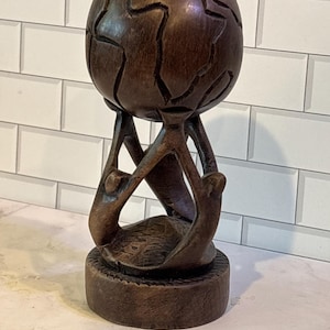 May include: A dark brown carved wooden sculpture featuring a spherical globe with etched continental outlines. The globe is supported by three stylized figures with elongated limbs that rise from a circular base with a textured edge.