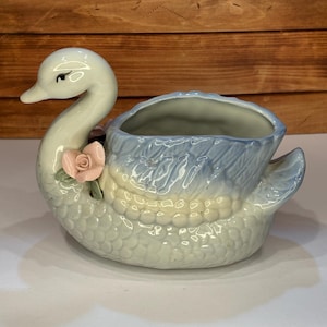 May include: A ceramic swan planter with a light blue and white color scheme. The swan has a long neck and a pink rose accent. The planter is designed to hold small plants or flowers, adding a touch of elegance to any space.