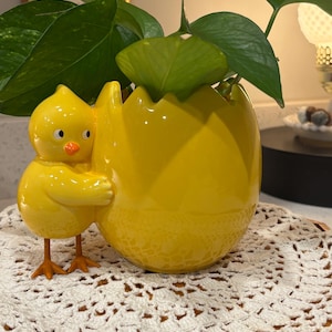 May include: A yellow ceramic Easter-themed planter shaped like a cracked egg, with a chick figurine attached. The planter holds green leafy plants and sits on a white crocheted doily. The chick has an orange beak and feet.