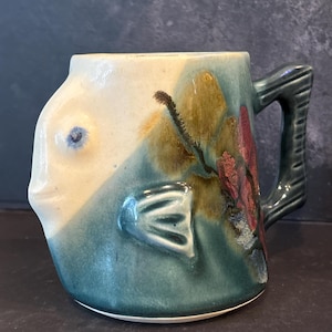 May include: A ceramic mug shaped like a fish with a cream-colored head and a teal-colored body. The mug features a raised fin on the side, a textured handle, and a painted floral design in shades of brown and red on the side of the fish body.