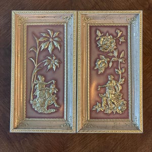 May include: Two framed wall art pieces. Each features a gold relief design on a mauve background. One depicts figures with palm leaves, the other roses and figures. The frames are gold and off-white.