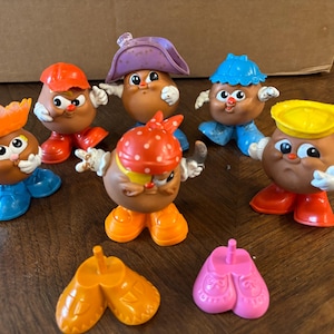 May include: A collection of six small potato-shaped plastic figures with various hats, shoes, and facial features, including a pirate-themed figure with an eye patch, plus two extra pairs of plastic shoes in orange and pink colors.
