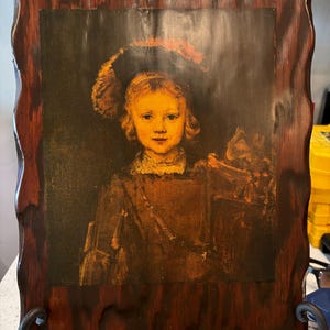 May include: A framed portrait of a young person in a dark outfit and hat. The portrait is set within a dark, wavy-edged wooden frame. The artwork features warm tones of brown, orange, and yellow, creating a vintage aesthetic. The frame is supported by a black metal stand.
