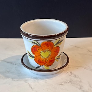 May include: A white ceramic flower pot with a matching saucer, featuring a hand-painted orange flower design with yellow centers and dark green leaves. The rim of the pot and the edge of the saucer are accented with a thin, dark brown painted line.