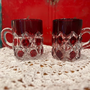 May include: Two small, ruby red glass mugs with clear handles and diamond-shaped cut crystal designs. The mugs sit on a white, crocheted doily. The background is a blurred red and turquoise.