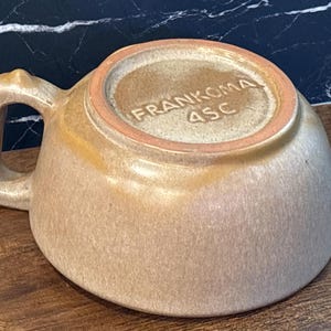 May include: A tan ceramic mug with a single handle, shown upside down. The base features the embossed text FRANKOMA 4SC. The glaze has a mottled, textured appearance with lighter and darker beige tones across the surface of the cup.