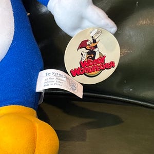 May include: A plush Woody Woodpecker toy, featuring a blue body, yellow feet, and white accents. The toy has a tag with the Woody Woodpecker logo and the text "Toy Network". The toy is made of polyester fiber.