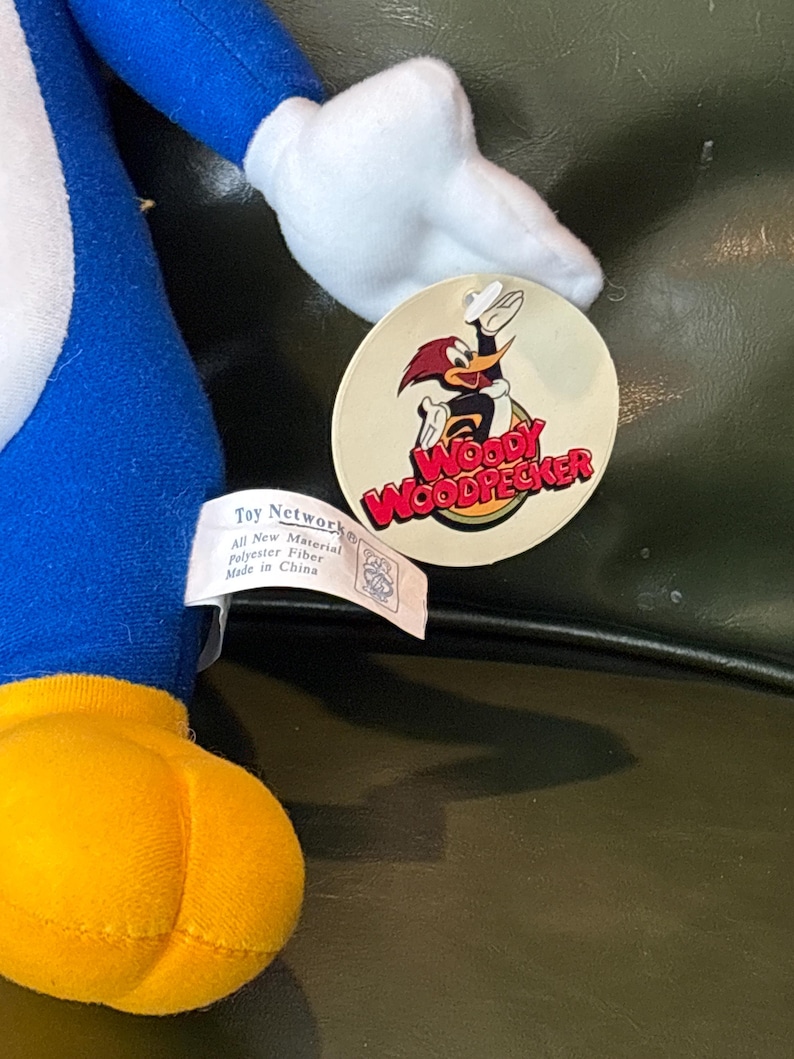May include: A plush Woody Woodpecker toy, featuring a blue body, yellow feet, and white accents. The toy has a tag with the Woody Woodpecker logo and the text "Toy Network". The toy is made of polyester fiber.