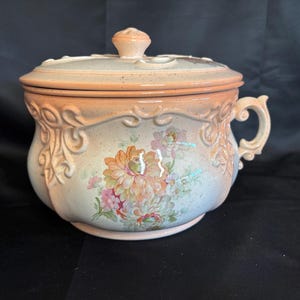 May include: A vintage ceramic chamber pot with a lid. The pot is painted with floral designs in shades of pink, orange, and green. It has a handle and a decorative rim. The lid is topped with a small knob. The pot is a light peach color.