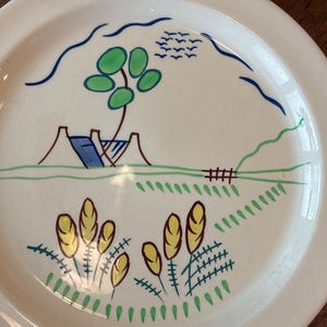May include: A round, off-white ceramic plate featuring a hand-painted landscape design. The illustration includes a green tree, a small blue and brown structure, a brown fence, yellow wheat stalks, and a flock of birds in the sky, all outlined in blue.