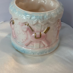May include: A ceramic carousel-themed planter in shades of pink, white, and blue. The planter features a carousel design with pink horses, gold accents, and star details. The top and bottom rims are light blue.