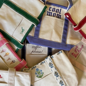 May include: Several off-white canvas tote bags with coloured trim in green, red, and purple. Some bags have embroidered text, including "cool mom", "yo", and "coffee please!" Other bags feature ice cream cones and floral designs.