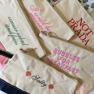 May include: Several canvas zipper pouches with embroidered text and designs. The pouches display phrases such as "I could be meaner" and "Salty". The pouches have a neutral color with colorful embroidery.