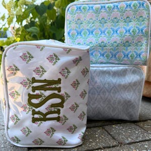 Op de afbeelding: Drie rechthoekige toilettassen met witte biezen. Eén tas heeft een witte basis met een herhalend roze en groen bloemenpatroon en een olijfgroen geborduurd monogram. Twee andere tassen tonen blauwe en groene bloemen- of geometrische patronen.