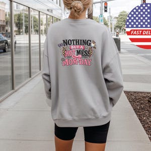 Peut inclure: Un sweat-shirt gris clair avec l'inscription "NOTHING BUT A HOT MESS MOM-DAY" dans un design coloré. Le motif comprend un drapeau à damier, un papillon et des accents roses. Le sweat-shirt est porté avec un short noir.
