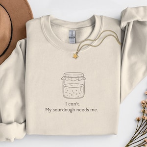 Könnte beinhalten: Cremefarbenes Sweatshirt mit Rundhalsausschnitt und langen Ärmeln. Auf der Vorderseite befindet sich eine Strichzeichnung eines Sauerteigglases mit dem Text "I can't. My sourdough needs me." Ein goldener Sternanhänger hängt an einer zarten Kette.