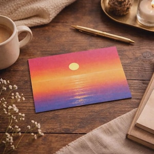 May include: A greeting card featuring a sunset scene with a yellow sun over a body of water, transitioning from pink to orange and purple hues. The card is on a wooden surface with a cup of coffee, a pen, and a candle.