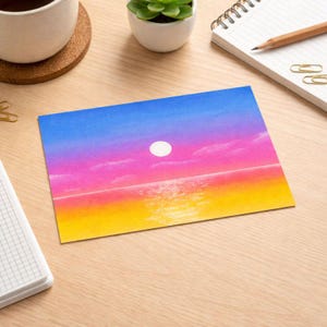 Könnte beinhalten: Ein rechteckiger Kunstdruck mit einer Sonnenuntergangsszene. Der Himmel verläuft von Blau über Rosa nach Gelb, mit einer weißen Sonne. Das Meer spiegelt die Farben des Sonnenuntergangs wider. Der Druck liegt auf einer Holzoberfläche, mit einer Kaffeetasse, einer Pflanze und einem Notizblock in der Nähe.