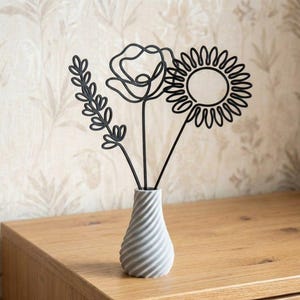 May include: A grey ceramic vase with a spiral design holds three black metal floral sculptures: a sprig, a poppy, and a sunflower. The vase sits on a wooden surface against a patterned wallpaper background.