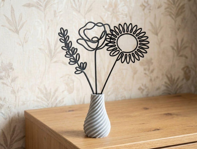 May include: A grey ceramic vase with a spiral design holds three black metal floral sculptures: a sprig, a poppy, and a sunflower. The vase sits on a wooden surface against a patterned wallpaper background.