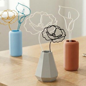 May include: A selection of wire art flowers displayed in vases. The flowers are crafted in gold, white, black, and teal wire. The vases are in blue, grey, and terracotta. The arrangement is set on a wooden surface.