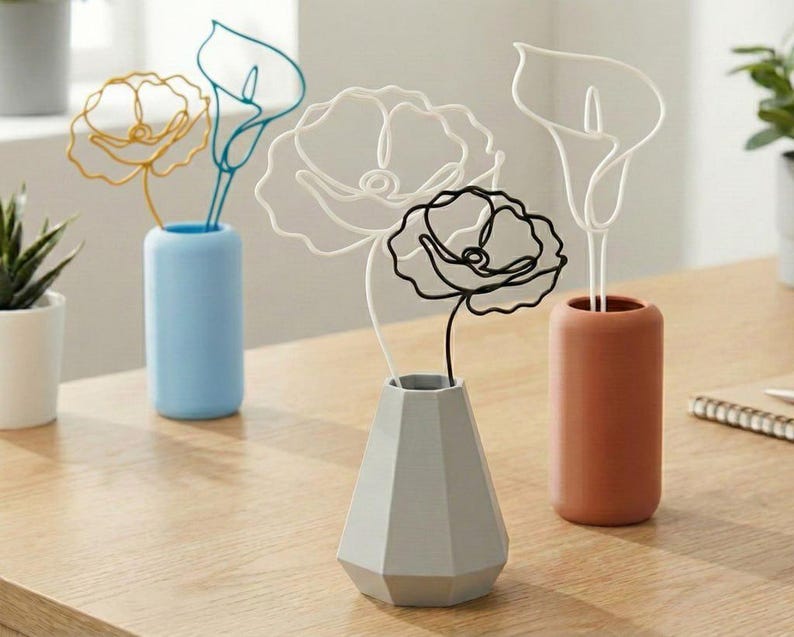 May include: A selection of wire art flowers displayed in vases. The flowers are crafted in gold, white, black, and teal wire. The vases are in blue, grey, and terracotta. The arrangement is set on a wooden surface.