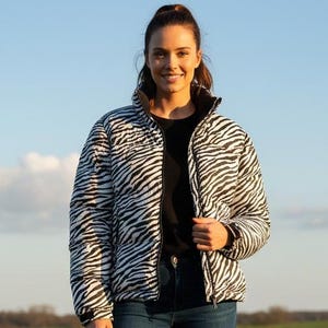 Puede incluir: Chaqueta acolchada con estampado de cebra en blanco y negro. La chaqueta se lleva sobre una camiseta negra y se combina con vaqueros azules y botas marrones. La persona está de pie en un campo de hierba con una valla de madera al fondo.