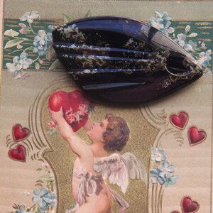 May include: A vintage Valentine's Day card featuring a cherub holding a red heart and a black and white patterned brooch. The card has a gold border and is decorated with flowers and hearts.