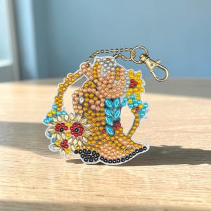 Könnte beinhalten: Ein dekorativer Schlüsselanhänger mit einer Frau in gelbem Kleid und einem Blumenstrauß, verziert mit bunten Strasssteinen. Der Schlüsselanhänger hat eine goldfarbene Kette und einen Verschluss, vor hellem Hintergrund.