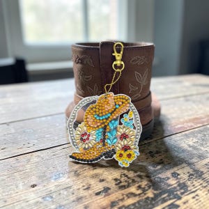 Könnte beinhalten: Ein dekorativer Schlüsselanhänger mit einem Strass-besetzten Cowboy-Stiefel-Design und einem goldfarbenen Verschluss. Der Stiefel-Anhänger hat ein gelbes, blaues und rotes Farbschema und hängt vor einem braunen Lederstiefel.